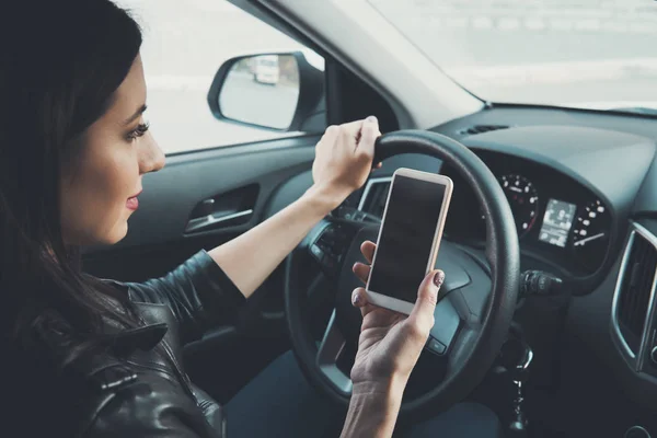 Mujer joven mirando a su teléfono inteligente mientras conduce el coche en el fondo de la ventana delantera. Conductora distraída. Concepto de conducción peligrosa. Mujer conduciendo y sosteniendo el teléfono celular con pantalla en blanco — Foto de Stock
