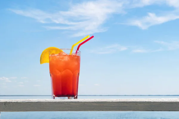 Bocal de cóctel afrutado en una mesa de playa. vaso de jugo de bayas rojas con una pajita y una rebanada de naranja sobre una mesa de madera en un bar junto al mar. en el fondo de un hermoso paisaje marino . — Foto de Stock