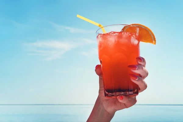 Mädchen hält einen roten Cocktail mit einer Orangenscheibe in der Hand. Orangensaft in Frauenhand am Strand mit Meereshintergrund — Stockfoto
