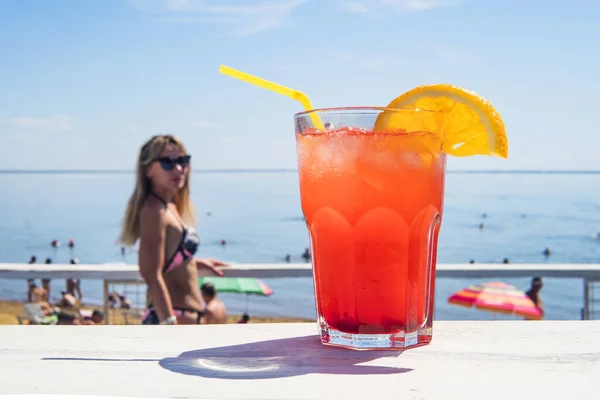 Um coquetel laranja colorido na mesa contra o pano de fundo de muitos turistas e meninas turistas. Sumo de cor vermelha em um copo na mesa branca contra o mar e o céu azul. lazer, atividade recreativa — Fotografia de Stock