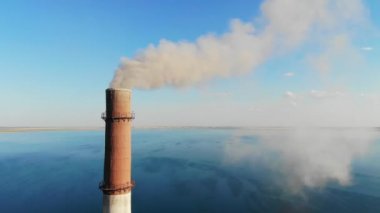 Gray smoke comes out of a large brick chimney in the factory against a clear blue sky and a large lake. 4K video. Environmental pollution. bad ecology, modern problems. smog