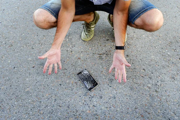 Парень уронил свой мобильник на асфальт и сломал его. Разочарованный человек со сломанным телефоном. мужчина сидит и держит сломанный смартфон с треснувшим экраном — стоковое фото