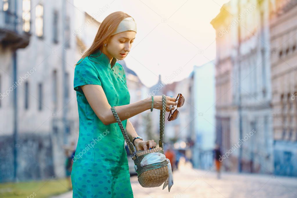 Young beautiful fashionable lady wearing green dress searching for something in her hand bag on a street of the old city. Female fashion. City lifestyle