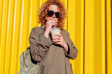 Hipster sarı duvara kahve içme sırt çantası olan genç kadın. Şık yaz gezgin açık havada rahatlatıcı güneş gözlüğü