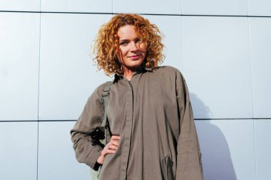 Kırmızı kıvırcık saçlı ayakta gri duvar karşı olan modern kadın. Açık havada şık yaz kıyafeti giymiş hipster modeli