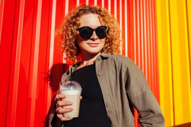 Genç ve güzel kadın kahve kırmızı duvara tutan kıvırcık kırmızı saçlı. Şık yaz kız gülümseyerek ve açık havada rahatlatıcı güneş gözlüğü