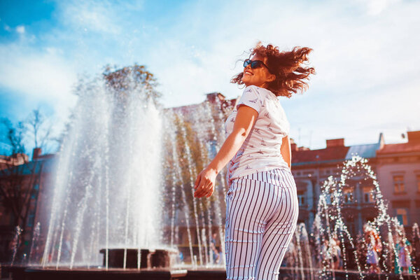 Happy young woman running and dancing by fountain on summer street. Hipster teen girl having fun