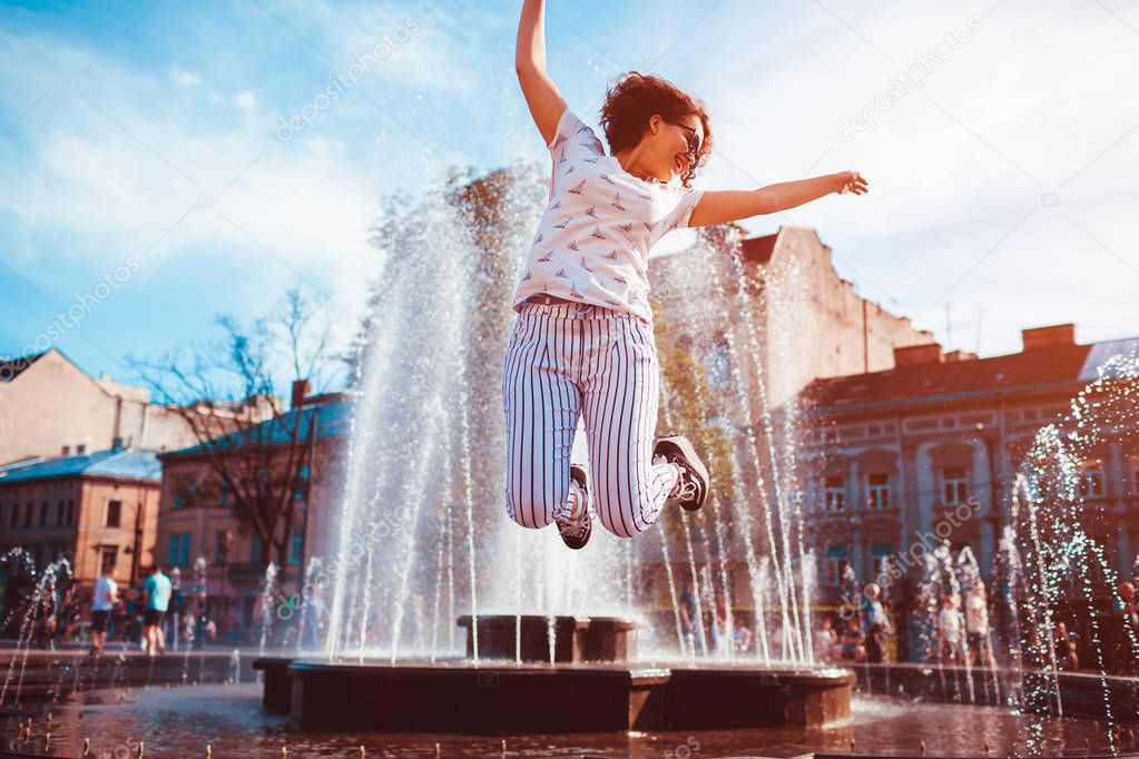 Happy young woman jumping by fountain on summer street. Hipster teen girl having fun