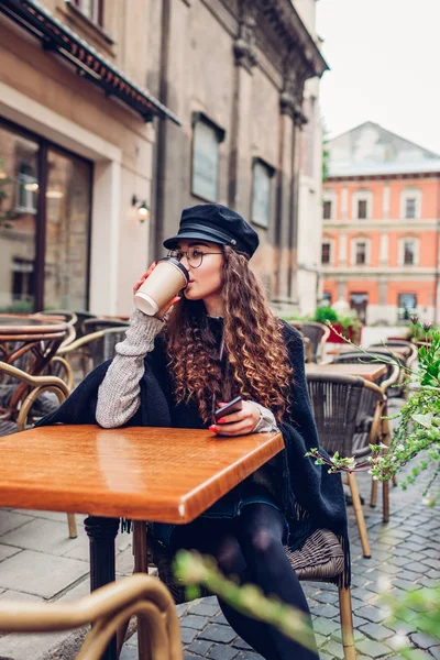 スタイリッシュな若い女性がスマート フォンを使用している間 屋外カフェでコーヒーを飲んでいます 低温の街の女子大生の美容ファッション ポートレート — ストック写真