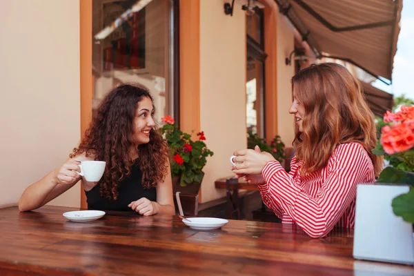 Happy female friends having coffee in outdoor cafe in summer on the street. Women chatting and chilling in the city