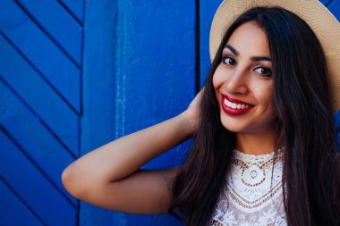 Mutlu Orta Doğulu kız gülümseyerek ve kameraya bakıyor. Açık mavi zemin üzerine yaz şapka giyen genç kadın portresi