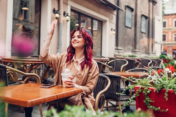 Beautiful young woman waving at friends while having coffee in outdoor cafe. Meeting in the city center for coffee break.