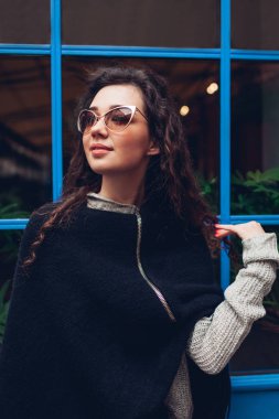 Mavi pencere karşı açık havada poz şık genç kadın. Şık kıyafet. Kıvırcık saçlı gülümseyen güzel model