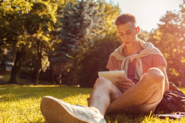 Yakışıklı üniversitelim Not defterini kampüs Park içinde yazmak. Mutlu erkek öğrenci açık havada bahar aylarında çim üzerinde oturan öğrenme. Eğitim