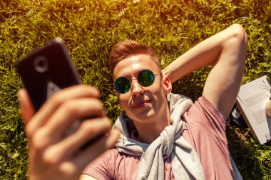 Yakışıklı üniversitelim bahar kampüs parkta soğutma. Gün batımında smartphone kullanarak çim ve alır selfie yatan mutlu erkek öğrenci