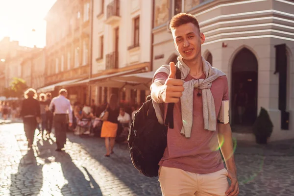 Yakışıklı üniversitelim şehir sokak yürüyüş. Mutlu erkek öğrenci bir kitap ve başparmak gösterilmesini sırt çantası ile. Eğitim kavramı