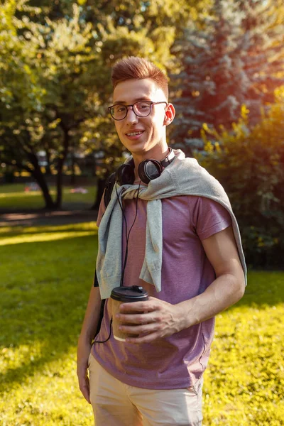 Handsome college guy chilling in spring campus park. Happy man student wears earphones and glasses and holds cup of coffee