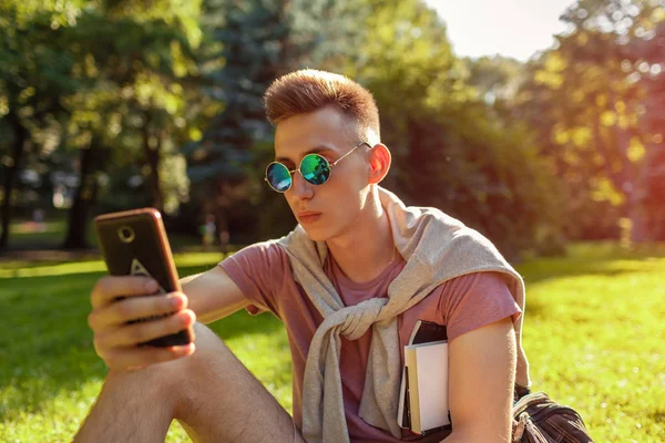 Yakışıklı üniversitelim smartphone kampüs Park içinde asılı. Mutlu erkek öğrenci açık havada bahar aylarında çim üzerinde oturan öğrenme