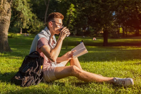 Handsome college man reading a book and drinking coffee in campus park. Happy guy student learning outdoors sitting on grass in spring. Education concept