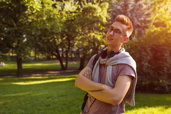 Handsome college guy chilling in spring campus park. Happy man student wears earphones and glasses and looks at camera
