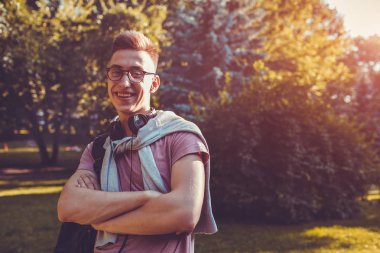 Yakışıklı üniversiteli bahar kampüs parkta soğutma. Mutlu bir adam öğrenci earphones ve gözlük takıyor ve kameraya benziyor