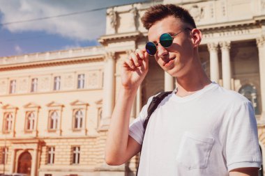 Yakışıklı üniversitelim gözlüklerini üniversite karşı denetleme. Mutlu erkek öğrenci kameraya gülümseyen sırt çantası ile. Eğitim