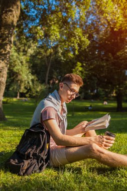 Bir kitap okuma ve kampüs parkta kahve içme yakışıklı üniversitelim. Mutlu erkek öğrenci açık havada bahar aylarında çim üzerinde oturan öğrenme. Eğitim kavramı