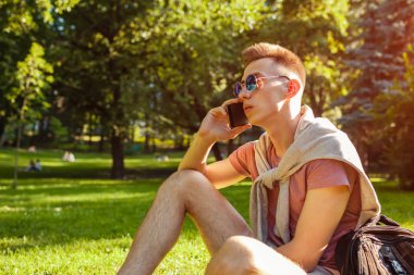 Yakışıklı üniversitelim smartphone kampüs Park içinde asılı. Mutlu erkek öğrenci açık havada bahar aylarında çim üzerinde oturan öğrenme