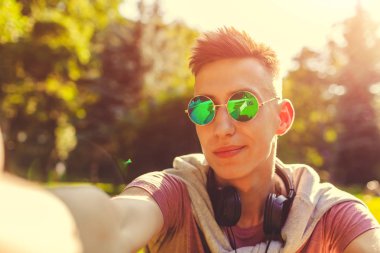 Yakışıklı üniversitelim bahar kampüs parkta soğutma. Gün batımında smartphone kullanarak güneş gözlüğü ve alır selfie mutlu erkek öğrenci