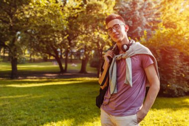 Yakışıklı üniversiteli bahar kampüs parkta soğutma. Mutlu bir adam öğrenci earphones ve gözlük takıyor ve kameraya benziyor