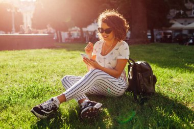 Akıllı telefon kullanarak ve kampüs Park müzik dinlemek güzel üniversiteli kız. Çimenlerin üzerinde oturan ve soğutma genç öğrenci