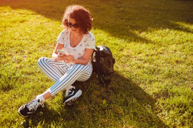 Akıllı telefon kullanarak ve kampüs Park müzik dinlemek güzel üniversiteli kız. Çimenlerin üzerinde oturan ve soğutma genç öğrenci