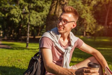 Bir kitap okuma ve kampüs parkta kahve içme yakışıklı üniversitelim. Mutlu erkek öğrenci açık havada bahar aylarında çim üzerinde oturan öğrenme. Eğitim kavramı