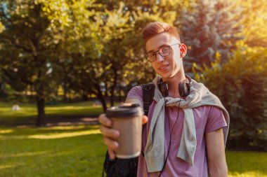 Yakışıklı üniversiteli bahar kampüs parkta soğutma. Kulaklık ve gözlük ve içme kahve mutlu erkek öğrenci giyer