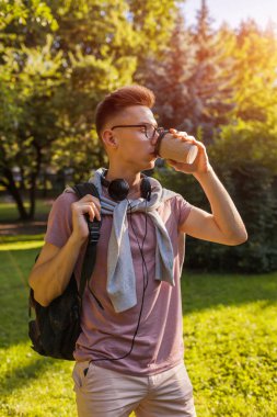 Yakışıklı üniversiteli bahar kampüs parkta soğutma. Kulaklık ve gözlük ve içme kahve mutlu erkek öğrenci giyer