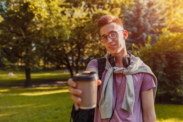 Handsome college guy chilling in spring campus park. Happy man student wears earphones and glasses and drinking coffee