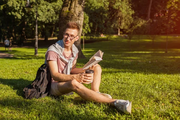 Handsome college man reading a book and drinking coffee in campus park. Happy guy student learning outdoors sitting on grass in spring. Education concept