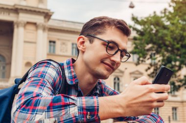 Yakışıklı üniversitelim bahar kampüs parkta soğutma. Mutlu erkek öğrenci ile smartphone kullanarak sırt çantası