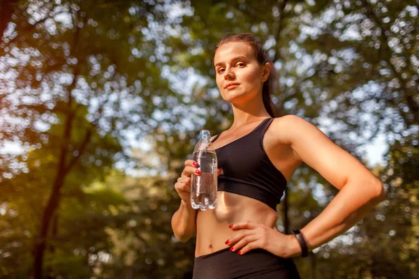 Young sportive woman drinking water after workout in summer park. Healthy way of life. Active lifestyle