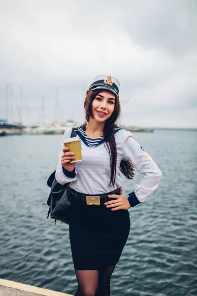 College woman student of Marine academy drinking coffee by sea wearing uniform. Girl walking in seaport of Odessa on pier