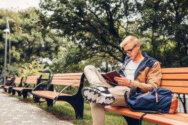 Üniversite öğrencisi sonbahar parkta bankta oturan yürüyüş kitap okuma sırt çantası ile. Adam açık havada eğitim