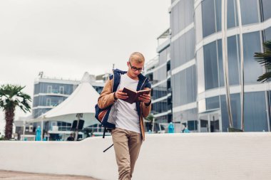 Üniversite öğrencisi tarafından modern otel tropik sahilde yürüyüş kitap okuma sırt çantası ile. Genç adam açık havada yağmurlu sonbahar gün eğitim