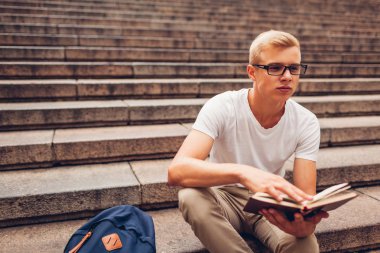 Merdivenlerde oturan ve gözlük tutan kitap okuma sırt çantası ile üniversite öğrencisi. Adam açık havada okuyor. Eğitim kavramı