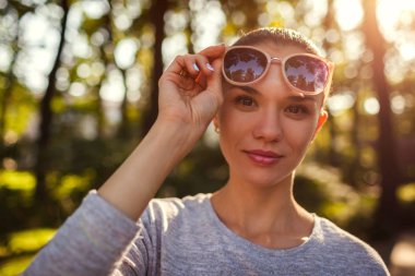 Gülümseyen genç kadın parkta onun güneş gözlüğü kapalı alarak. Doğal makyaj açık havada kameraya bakarak ile güzel kız