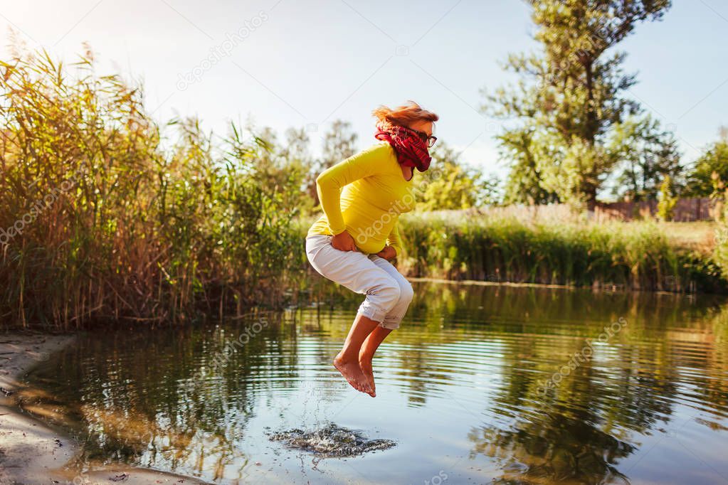 Middle-aged woman jumping on river bank on autumn day. Happy senior lady having fun walking in the forest. Feeling energetic and free