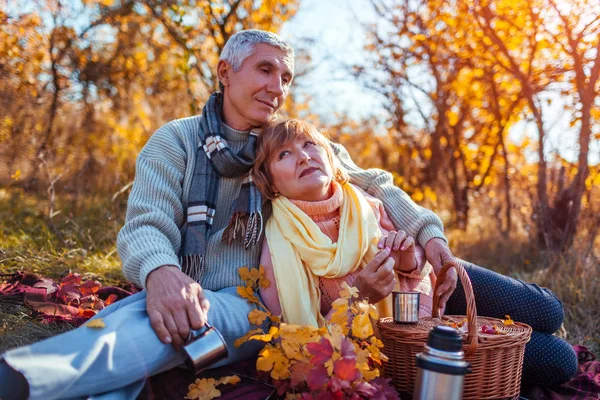 Senior couple having tea in autumn forest. Happy man and woman enjoying picnic and nature. Smiling man pouring tea out of thermos