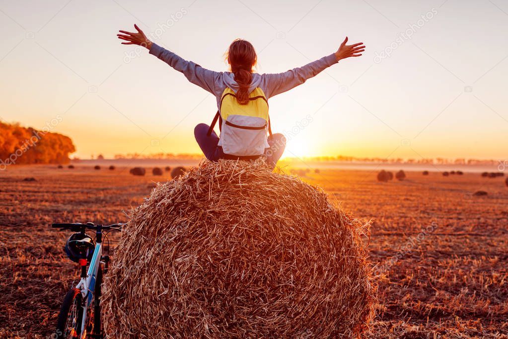 Young bicyclist sitting on haystack with raised and opened arms after a ride. Woman having rest in autumn field admiring view. Sportive lifestyle