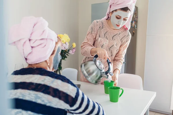 Молодая женщина со старшей матерью готовит чай в масках для лица. Женщины расслабляются и разговаривают на кухне — стоковое фото