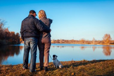 Sonbahar Park nehir tarafından üst düzey çift yürüyen pug köpek. Mutlu bir adam ve kadının açık havada pet ile vakit