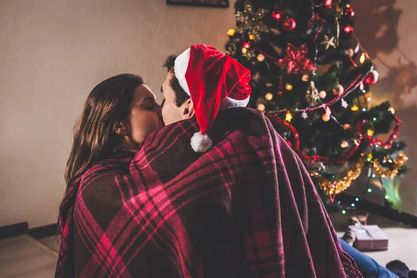 Couple in love sitting next to a Christmas tree, wearing Santa\'s hat and hugging. Young people celebrating New year at home at night